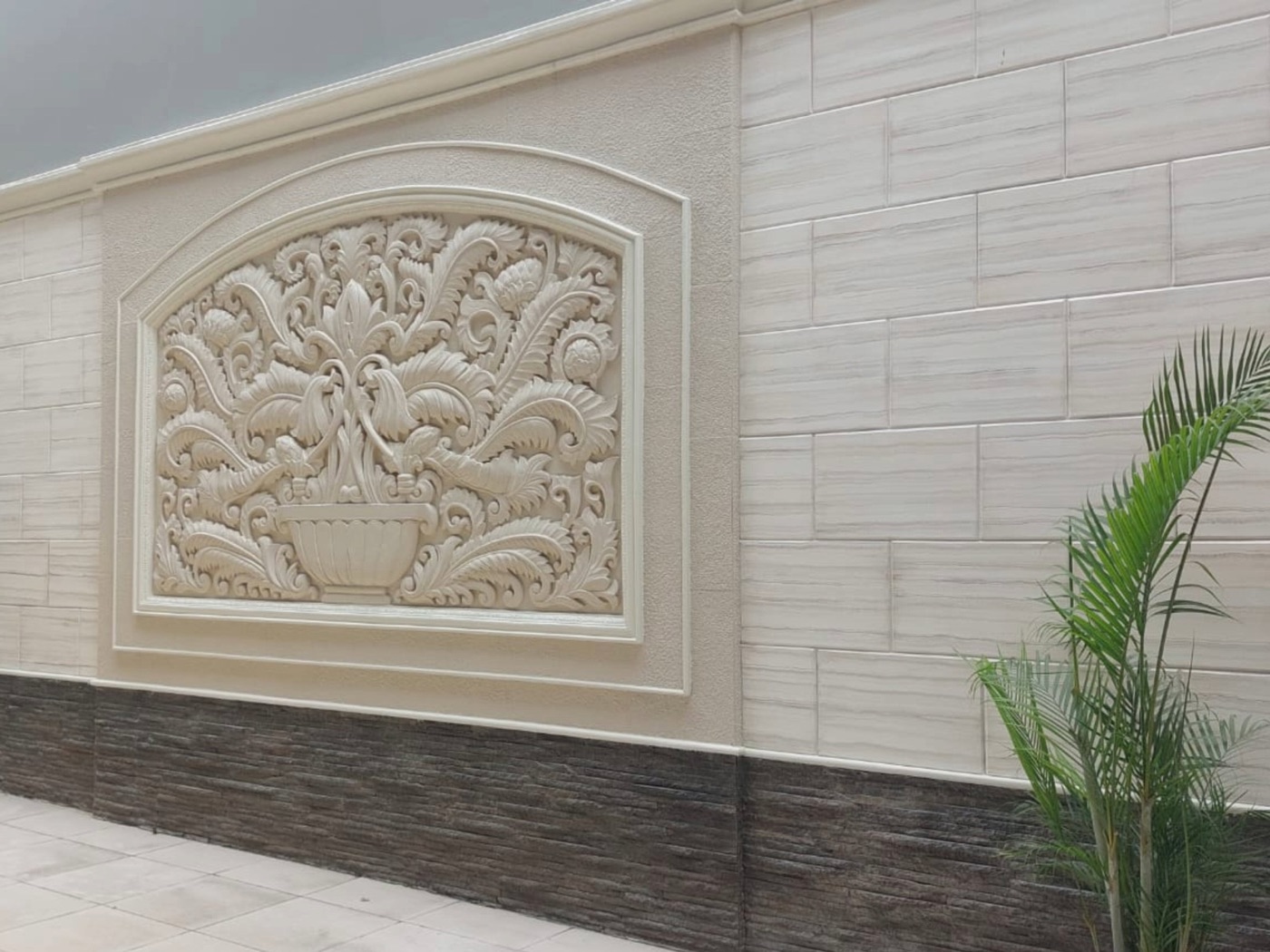 Carved relief panel set in a pale stone-clad wall with a palm in the corner